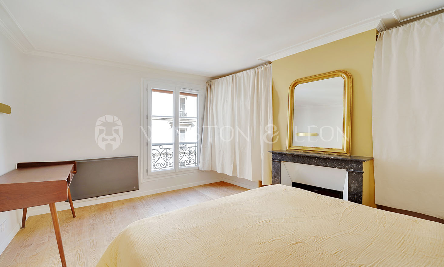 A minimalist bedroom in a premium apartment, featuring a large window and a yellow accent wall. A bed with matching blanket resides near a wooden desk, and to the right, theres a decorative fireplace adorned with a mirror. White curtains and light wooden flooring complete this Paris-inspired retreat.