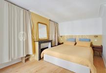 A cozy, furnished bedroom features a double bed draped in a beige duvet, flanked by premium wooden bedside tables. The yellow and white walls complement a black fireplace with a large mirror above, capturing hints of Parisian elegance. Beige curtains gently cover the windows.