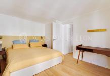 A minimalist bedroom in a furnished Paris apartment showcases a double bed with yellow bedding and two blue pillows. It features a wooden floor, a small desk with a chair on the right, and a white door. The light-colored walls are complemented by a wall-mounted fixture.