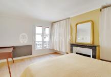 A minimalist bedroom in a premium apartment, featuring a large window and a yellow accent wall. A bed with matching blanket resides near a wooden desk, and to the right, theres a decorative fireplace adorned with a mirror. White curtains and light wooden flooring complete this Paris-inspired retreat.