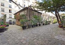 A quaint cobblestone courtyard featuring a rustic building with a wooden staircase and iron railings. This serene urban oasis, adorned with potted plants and surrounded by tall residential buildings, offers a premium atmosphere under a clear sky.