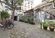 A quaint, cobblestone courtyard in Paris, bordered by potted plants and vines on trellis structures. Bicycles rest to the left, flanked by white buildings with windows, hinting at an urban setting. This premium spot feels like a beautifully furnished escape from the bustling city.