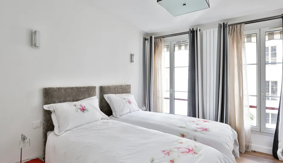 Bright, elegant bedroom with twin beds, floral bedding, upholstered headboards. Large windows and modern decor for luxury comfort.