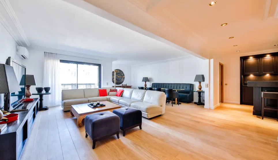 Spacious, bright luxury living room with light wood floors, modern sectional sofa, dining area, and integrated bar. High-end contemporary design.