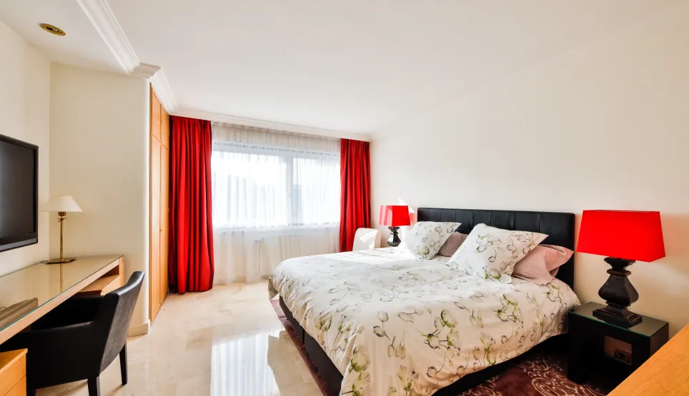 Stunning luxury bedroom with king-size bed, red curtains, marble floor, desk, and TV. Bright and modern design.