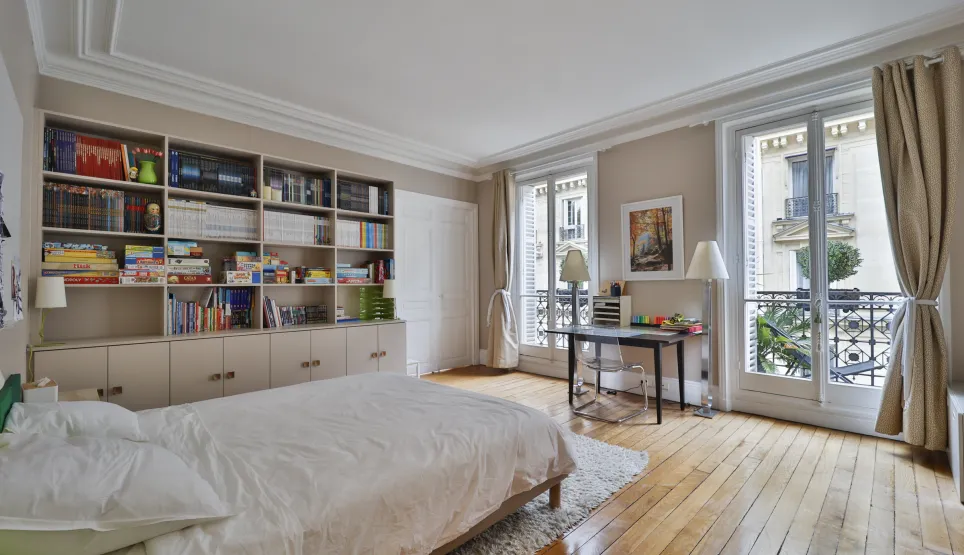 Bright Parisian bedroom with parquet floors, moldings, built-in library, and balconies. Elegance and comfort for a luxury apartment.