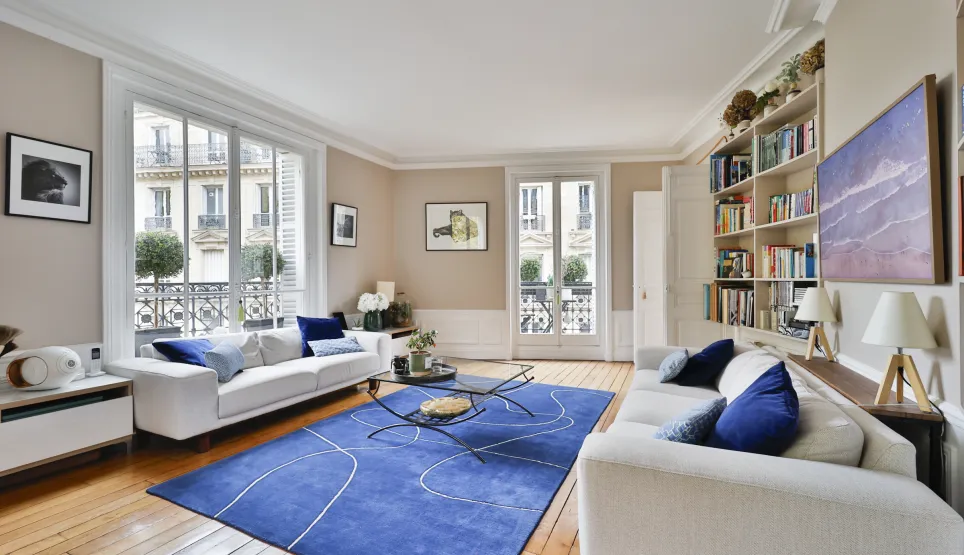 Bright luxury Parisian living room. White sofas, modern blue rug, hardwood floor. City view. Haussmannian elegance and comfort.