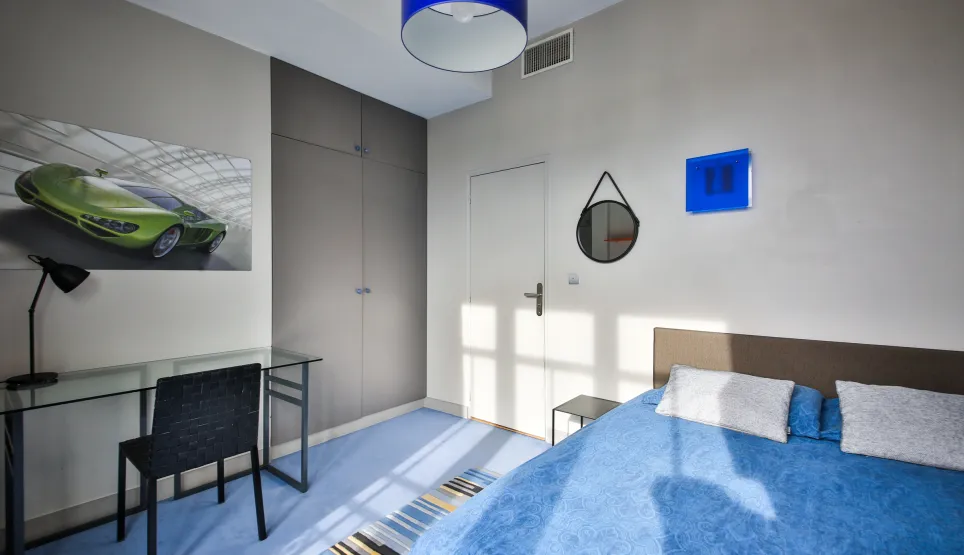 Modern luxury bedroom: bright blue bed, glass desk, built-in wardrobes, and sports car art. Elegant design for a contemporary living space.