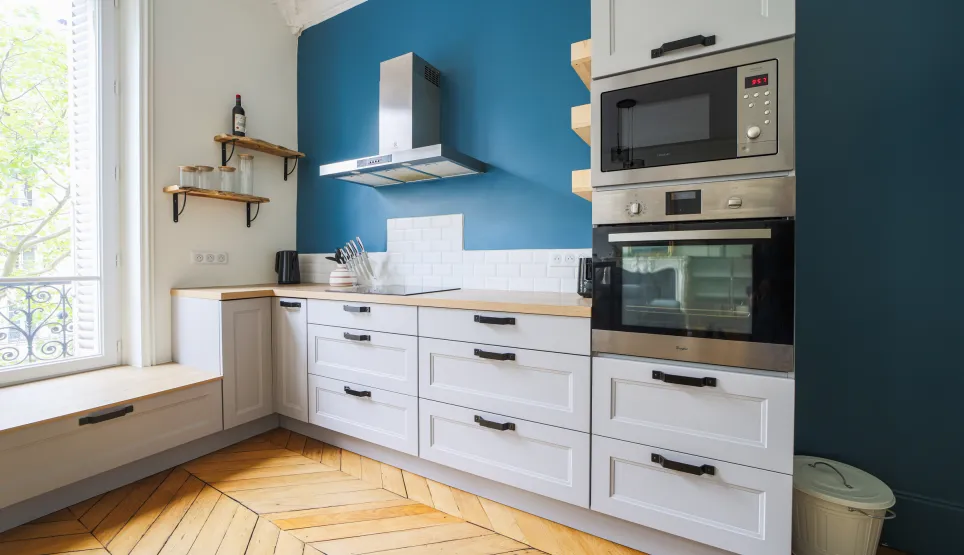 Bright modern kitchen with blue accent wall, herringbone parquet, white cabinetry & integrated appliances. Elegant Parisian apartment design.