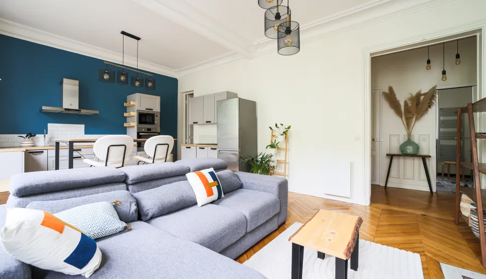 Modern open-plan living room and kitchen with grey sofa, blue accent wall, equipped kitchen, parquet flooring. Bright contemporary design.