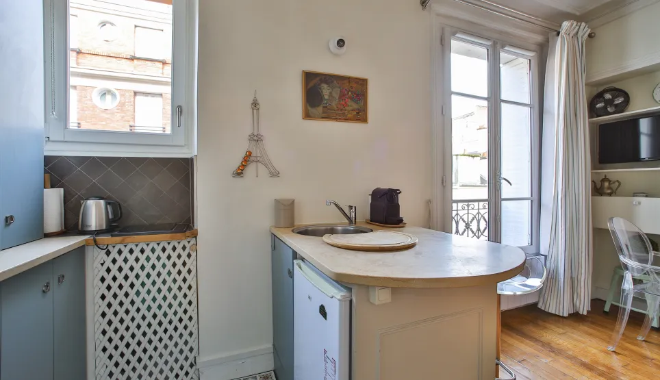 Optimized Parisian studio photo with integrated kitchen, bright window, hardwood floor, and Eiffel Tower decor. Chic, functional ambiance.