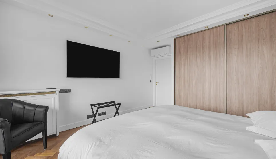 Modern luxury master bedroom featuring a double bed, wall-mounted TV, built-in wooden wardrobe, and design armchair. Elegant and comfortable.