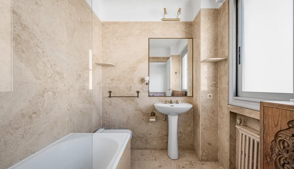 Elegant beige marble bathroom featuring a bathtub, pedestal sink, and large mirror. Bright, classic design for a luxury interior.