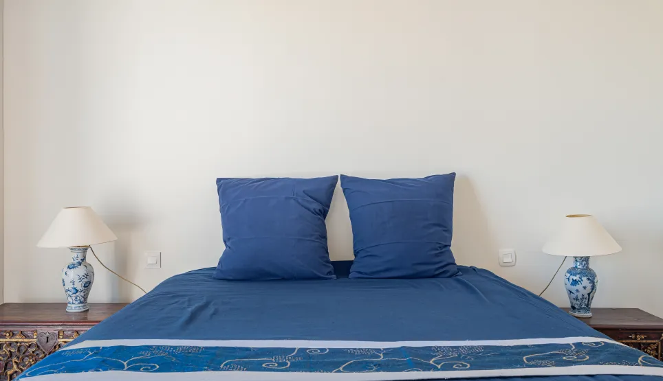 Elegant, comfortable bedroom featuring a deep blue bed, carved bedside tables, and porcelain lamps. Refined ambiance for a luxury property.