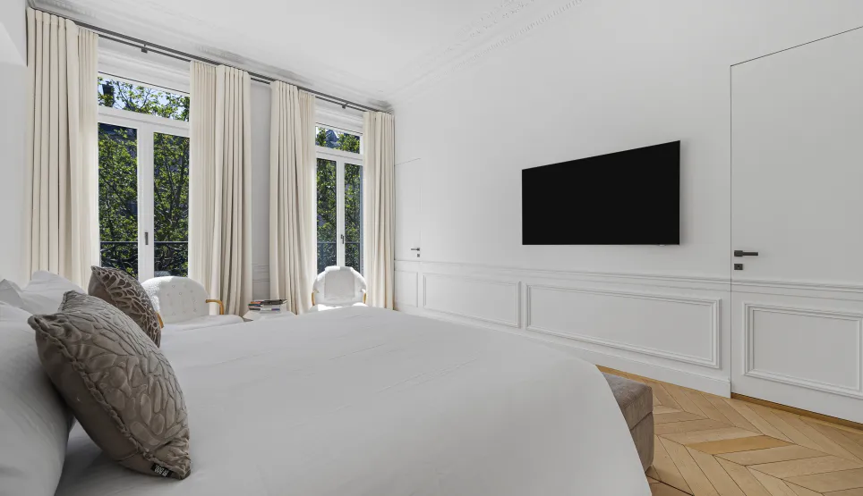 Bright, modern luxury bedroom. Large windows, natural light, herringbone parquet, sleek wall-mounted TV. Elegant and serene design.
