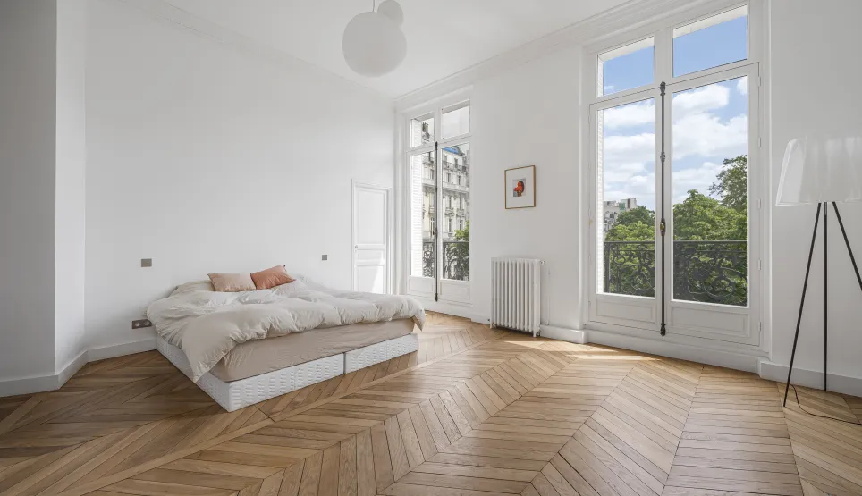 Bright Haussmannian apartment bedroom. Chevron parquet, high ceilings, large windows, green view. Elegant Parisian luxury.