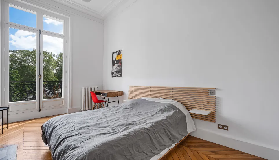 Bright bedroom with balcony overlooking greenery, herringbone parquet, and classic moldings. Modern design meets Parisian elegance for serene luxury.