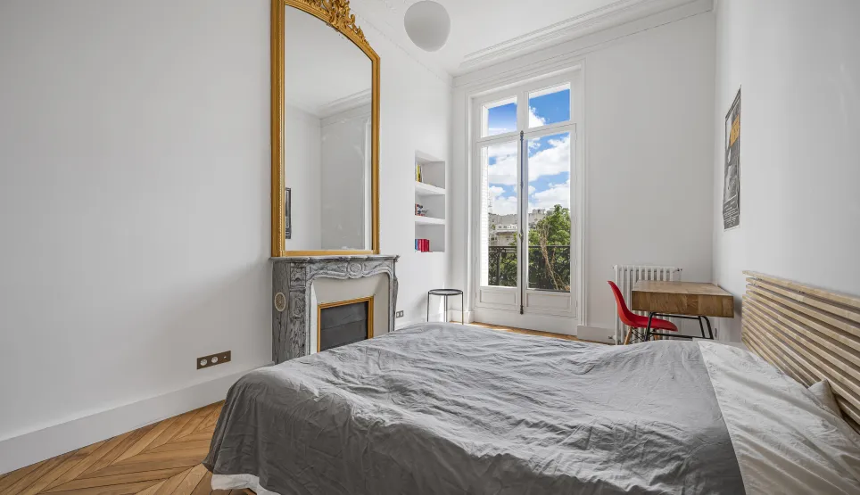 Bright bedroom with herringbone parquet, marble fireplace, ornate gold mirror, and open view. Haussmannian charm for a luxury apartment.
