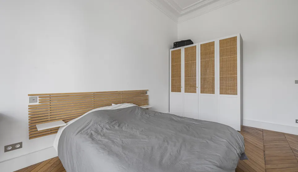 Sleek bedroom design: wooden headboard, rattan wardrobe, herringbone parquet. Serene and elegant ambiance for a luxury Parisian interior.