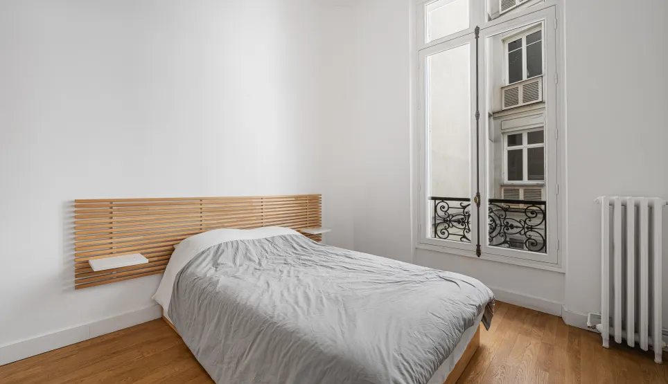 Bright, minimalist bedroom with hardwood floor, bespoke wooden headboard, and classic Parisian window. Luxury apartment.