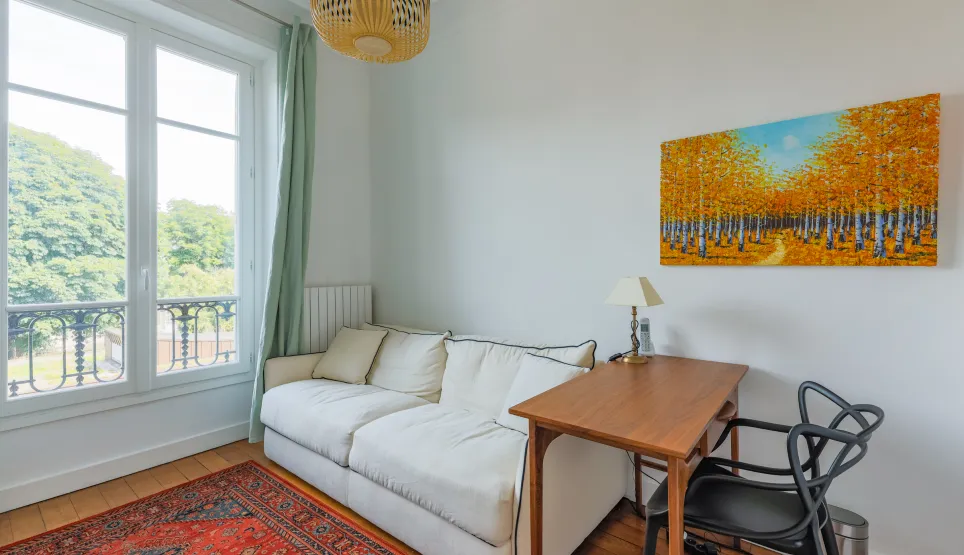 Bright room with green view, white sofa, wooden desk, and modern art. Serene, elegant ambiance for work or relaxation.