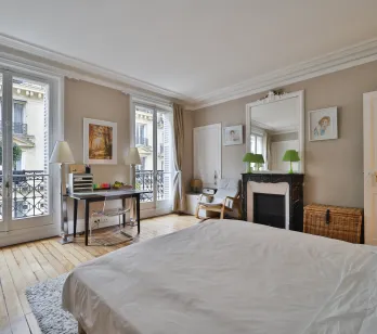 Luxury Haussmannian bedroom with balcony, hardwood floors, marble fireplace. Elegant suite, desk, moldings. Prestigious Parisian apartment.