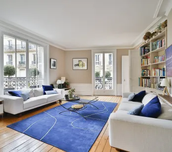 Bright luxury Parisian living room. White sofas, modern blue rug, hardwood floor. City view. Haussmannian elegance and comfort.