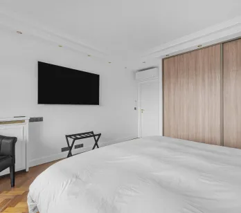 Modern luxury master bedroom featuring a double bed, wall-mounted TV, built-in wooden wardrobe, and design armchair. Elegant and comfortable.