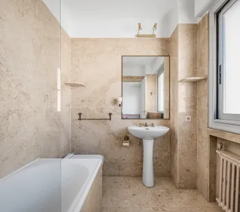 Elegant beige marble bathroom featuring a bathtub, pedestal sink, and large mirror. Bright, classic design for a luxury interior.