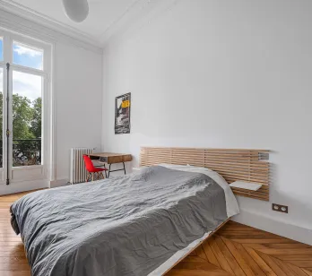 Bright bedroom with balcony overlooking greenery, herringbone parquet, and classic moldings. Modern design meets Parisian elegance for serene luxury.