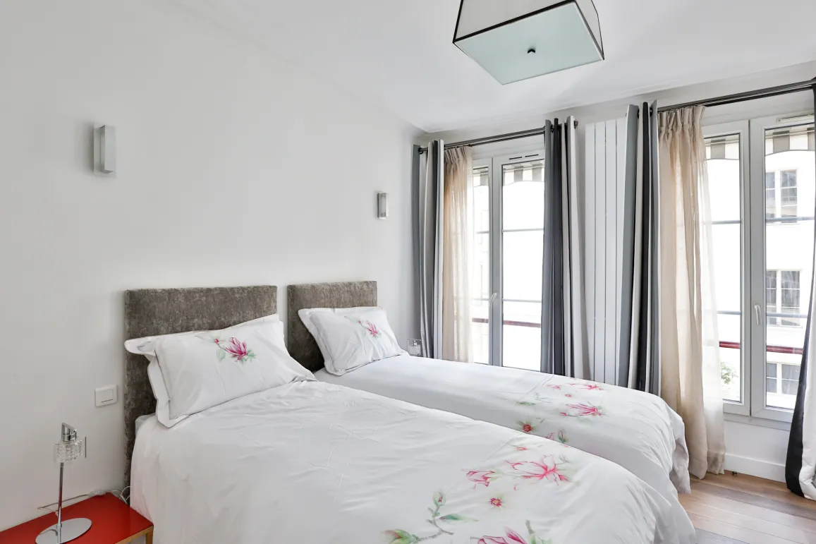 Bright, elegant bedroom with twin beds, floral bedding, upholstered headboards. Large windows and modern decor for luxury comfort.