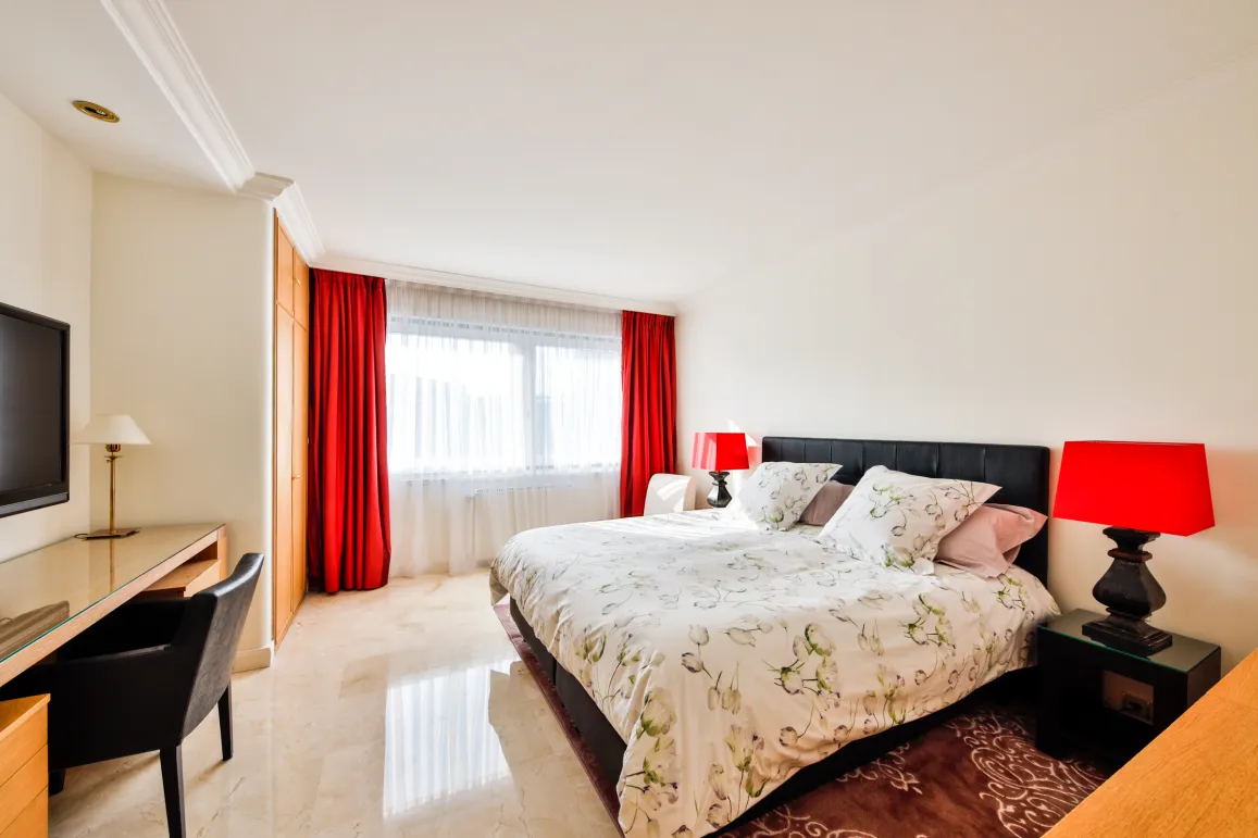 Stunning luxury bedroom with king-size bed, red curtains, marble floor, desk, and TV. Bright and modern design.