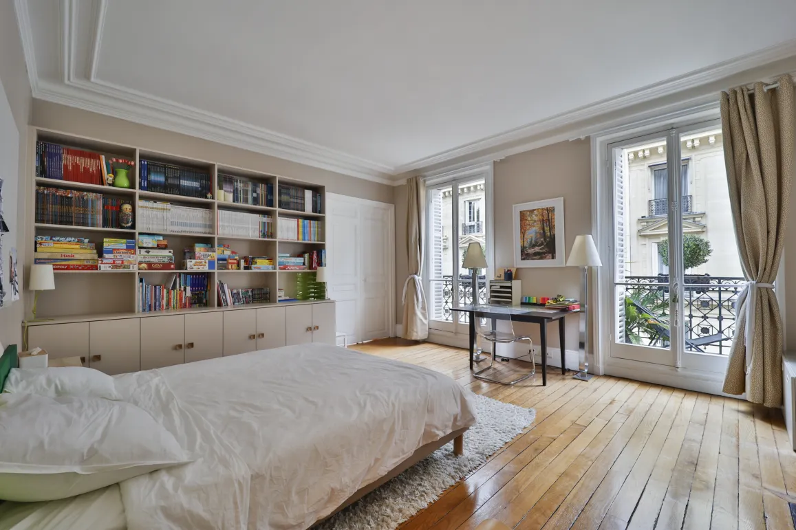 Bright Parisian bedroom with built-in library, hardwood floors, balcony, and city view. Ideal for luxury apartment.