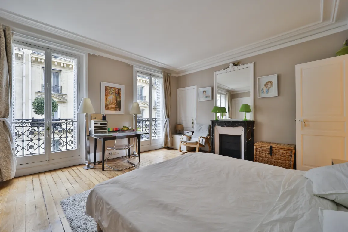 Luxury Haussmannian bedroom with balcony, hardwood floors, marble fireplace. Elegant suite, desk, moldings. Prestigious Parisian apartment.