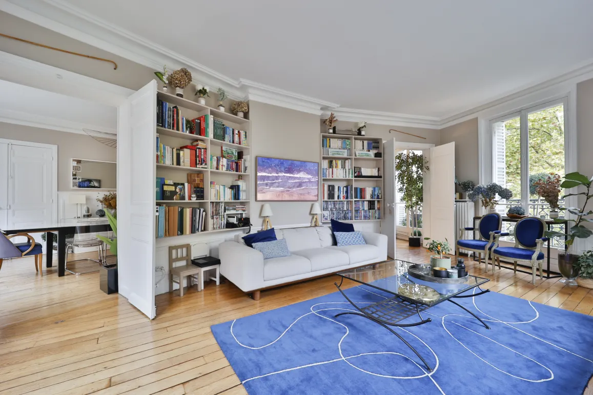 Expansive, bright living/dining room featuring hardwood floors, custom bookshelves, modern sofa, and lush green views. Luxury urban oasis.