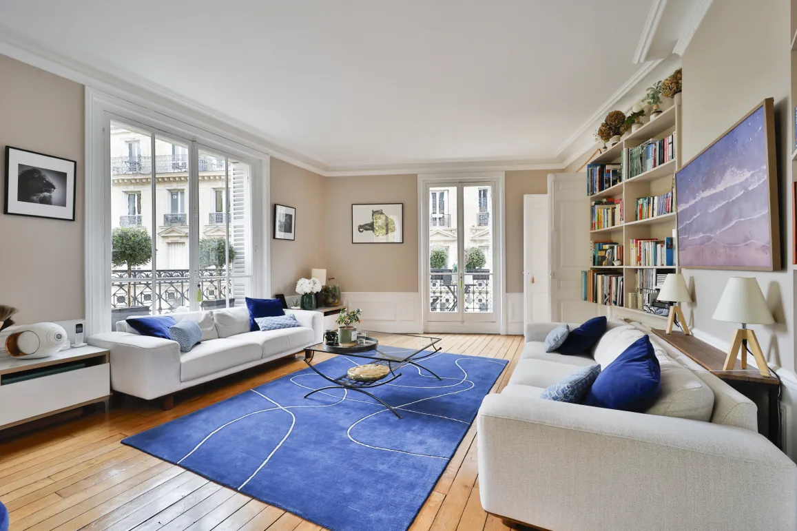 Bright luxury Parisian living room. White sofas, modern blue rug, hardwood floor. City view. Haussmannian elegance and comfort.