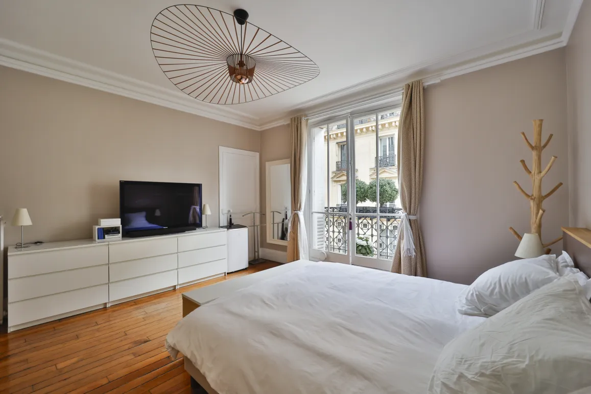 Bright, elegant bedroom with hardwood floors, large bed, TV, and balcony. Perfect for a luxury Parisian apartment. Modern design and comfort.