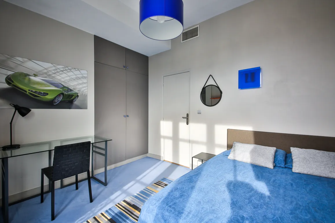 Modern luxury bedroom: bright blue bed, glass desk, built-in wardrobes, and sports car art. Elegant design for a contemporary living space.