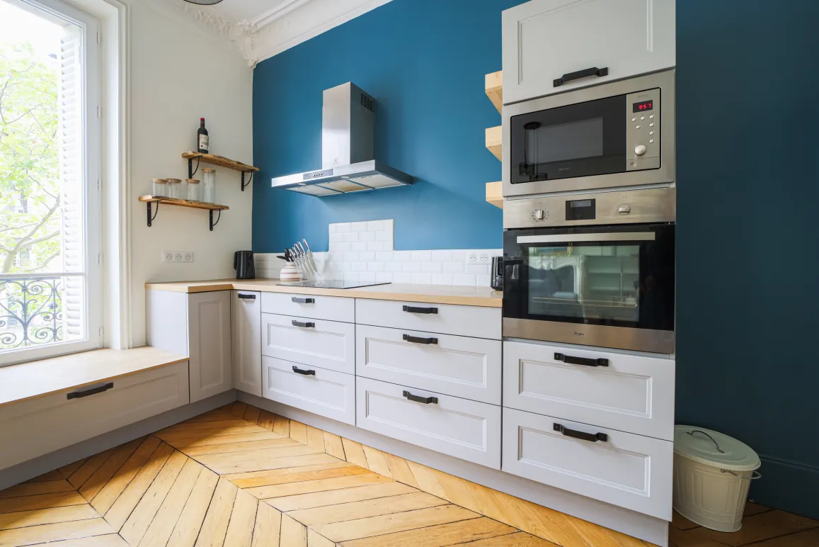 Bright modern kitchen with blue accent wall, herringbone parquet, white cabinetry & integrated appliances. Elegant Parisian apartment design.