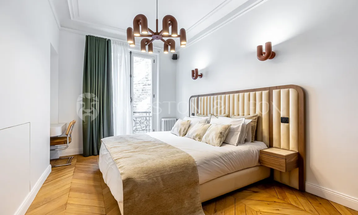 Modern luxury bedroom featuring a padded headboard, herringbone parquet, designer lighting, and balcony. Parisian elegance and comfort redefined.