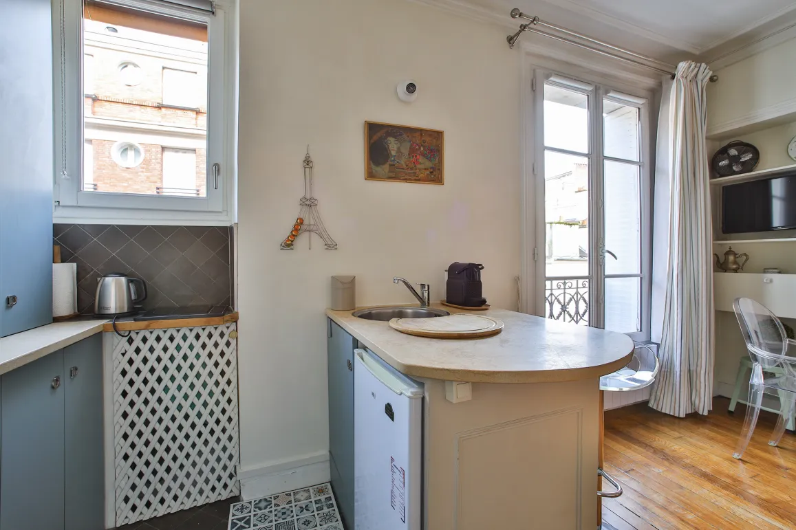 Optimized Parisian studio photo with integrated kitchen, bright window, hardwood floor, and Eiffel Tower decor. Chic, functional ambiance.