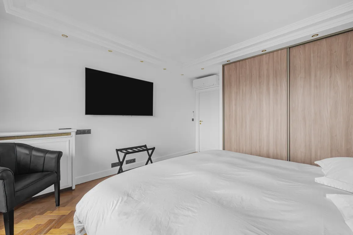 Modern luxury master bedroom featuring a double bed, wall-mounted TV, built-in wooden wardrobe, and design armchair. Elegant and comfortable.