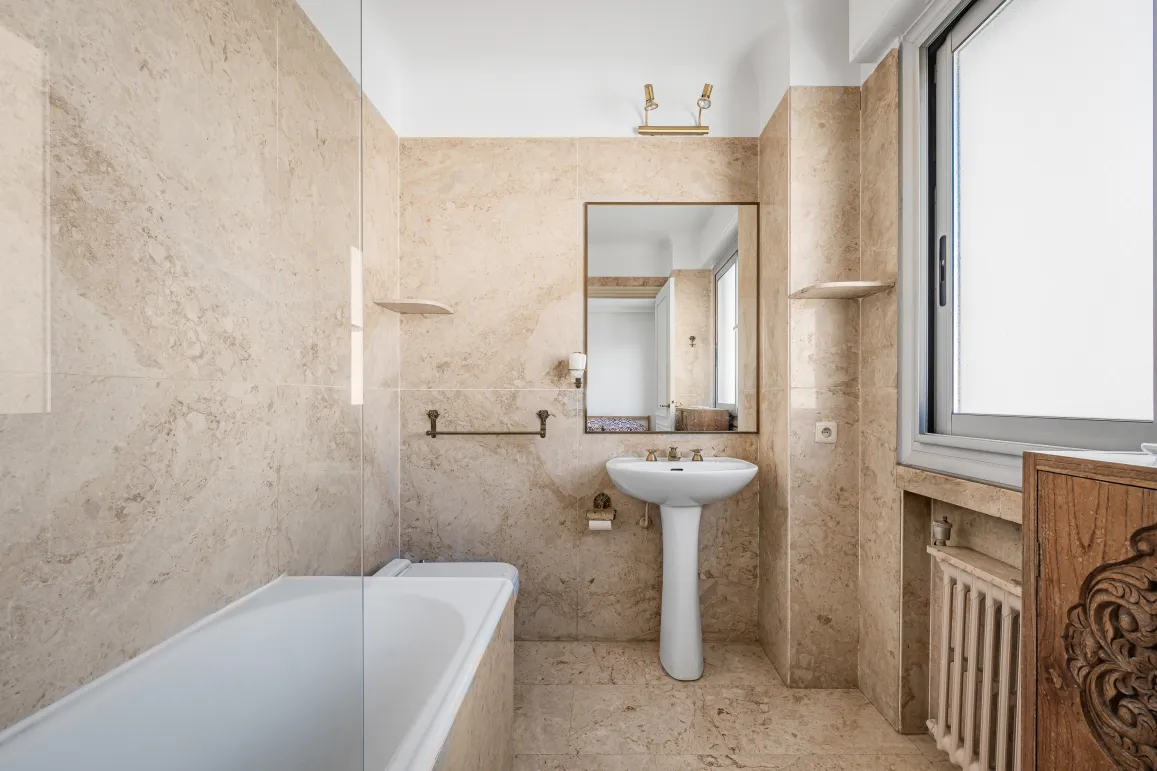 Elegant beige marble bathroom featuring a bathtub, pedestal sink, and large mirror. Bright, classic design for a luxury interior.
