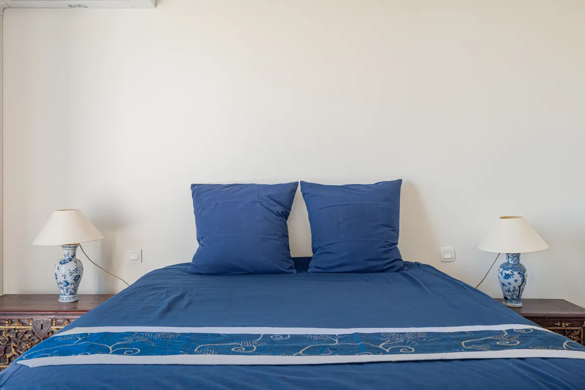 Elegant, comfortable bedroom featuring a deep blue bed, carved bedside tables, and porcelain lamps. Refined ambiance for a luxury property.