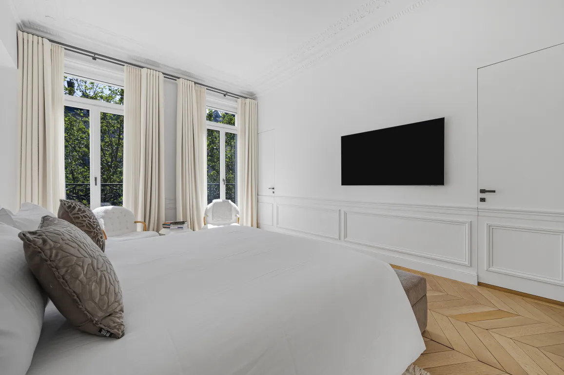 Bright, modern luxury bedroom. Large windows, natural light, herringbone parquet, sleek wall-mounted TV. Elegant and serene design.