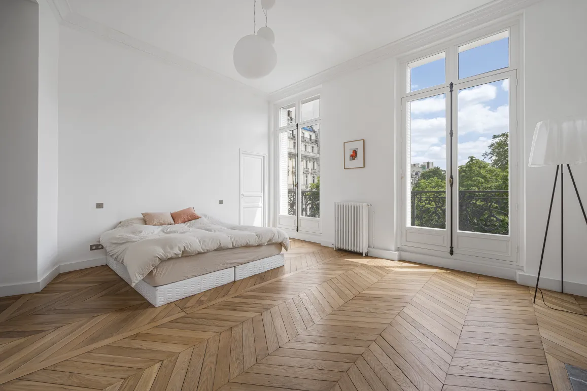 Bright Haussmannian apartment bedroom. Chevron parquet, high ceilings, large windows, green view. Elegant Parisian luxury.