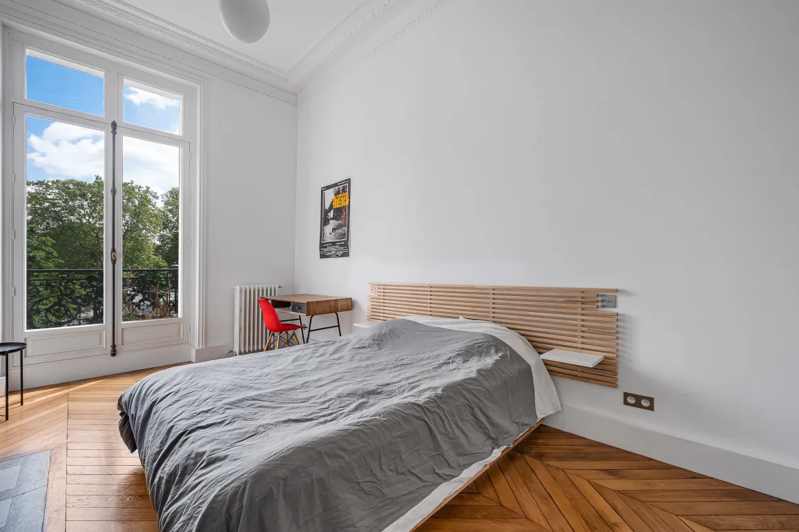 Bright bedroom with balcony overlooking greenery, herringbone parquet, and classic moldings. Modern design meets Parisian elegance for serene luxury.