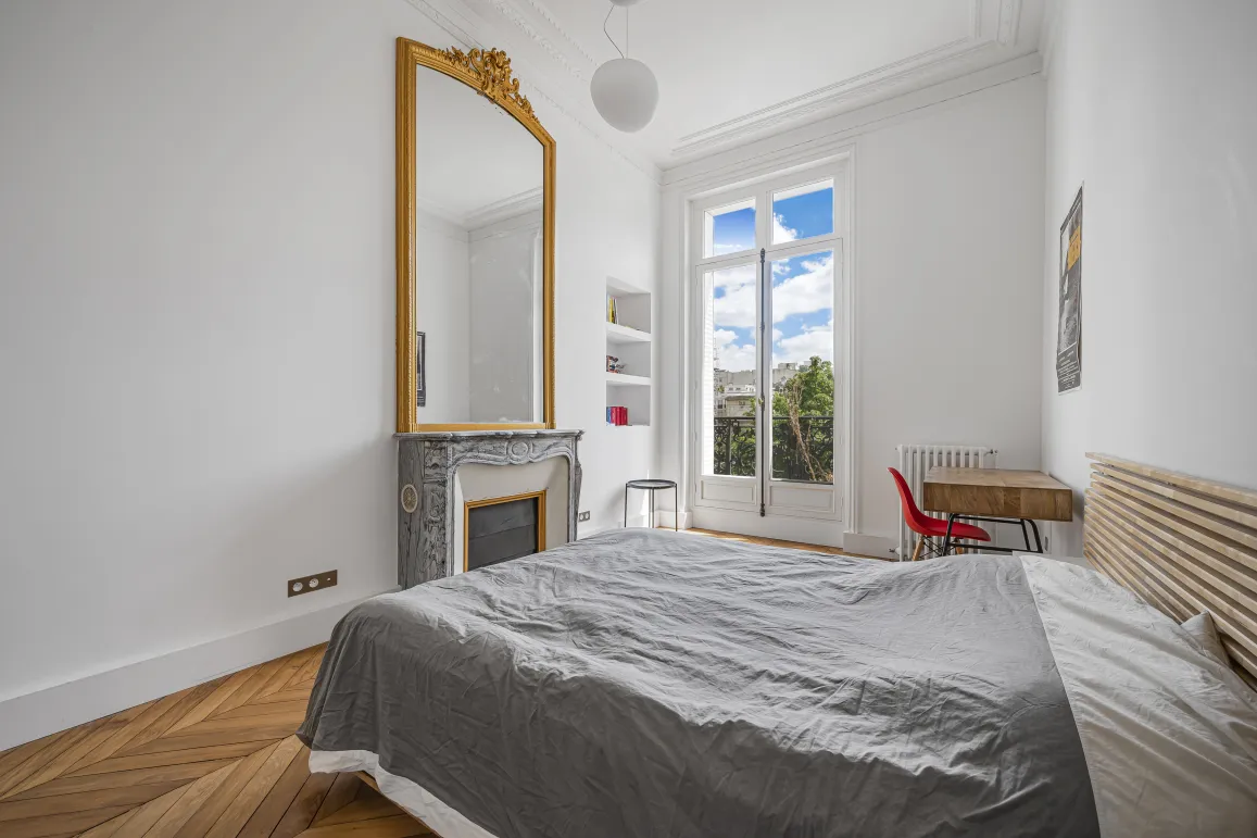 Bright bedroom with herringbone parquet, marble fireplace, ornate gold mirror, and open view. Haussmannian charm for a luxury apartment.