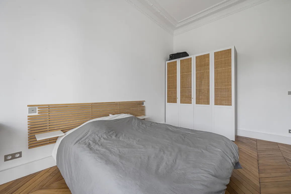 Sleek bedroom design: wooden headboard, rattan wardrobe, herringbone parquet. Serene and elegant ambiance for a luxury Parisian interior.