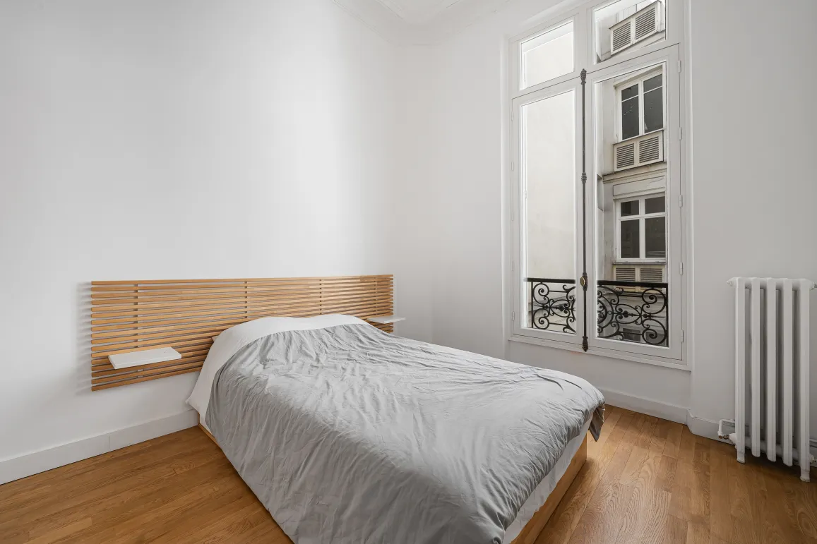 Bright, minimalist bedroom with hardwood floor, bespoke wooden headboard, and classic Parisian window. Luxury apartment.